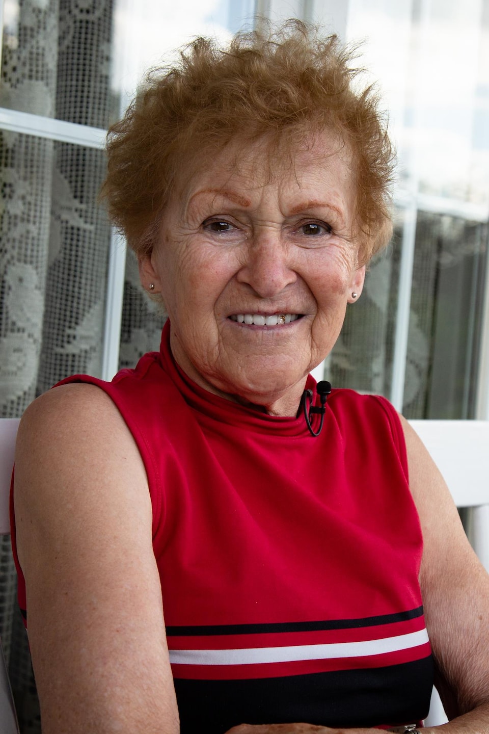 Une femme âgée pose pour la caméra, assise sur un banc à l'extérieur d'une maison. Elle a des cheveux roux courts et frisés.