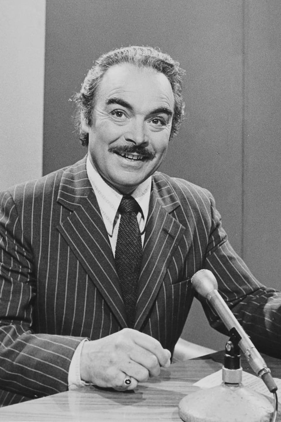 Henri Bergeron, carton à la main, animant assis à un bureau devant un micro sur pied.
