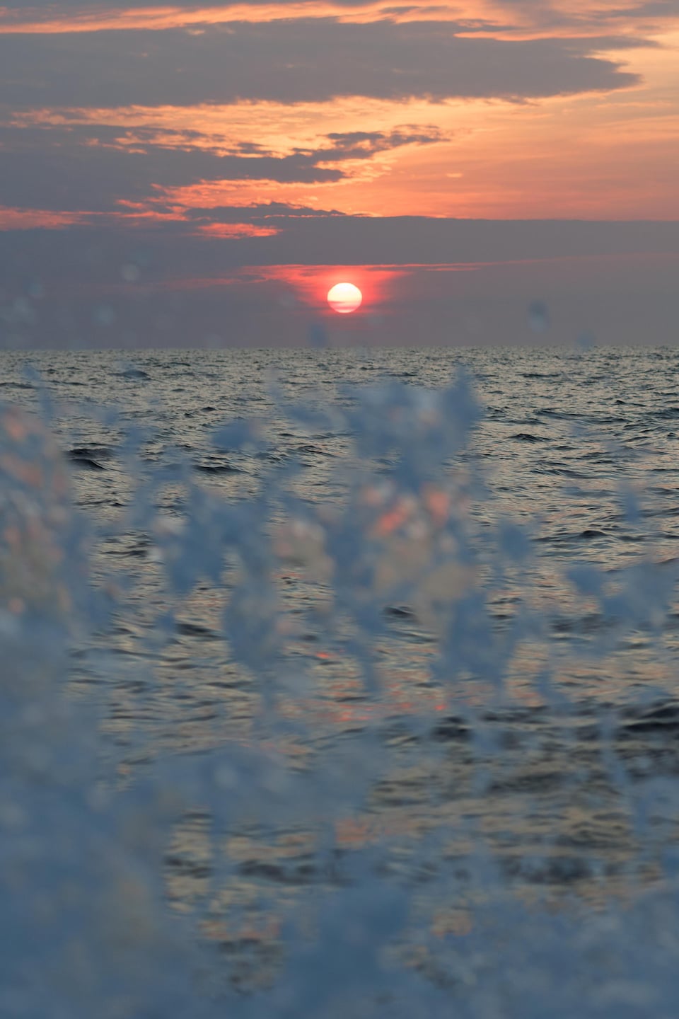 Des vagues créées par le bateau de Michaël Noël semblent atteindre le soleil d'un orange échelant et entouré de nuages bleus.