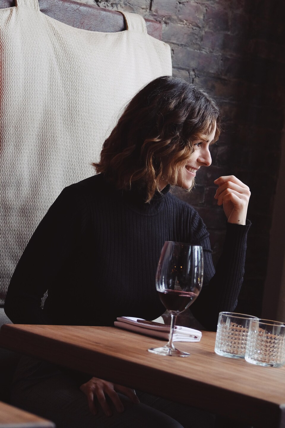 Caroline Beaulieu regarde au loin en rigolant. Elle est assise à une table de restaurant avec, en arrière-plan, des coussins et de la brique. Un verre de vin rouge est déposé sur sa table.