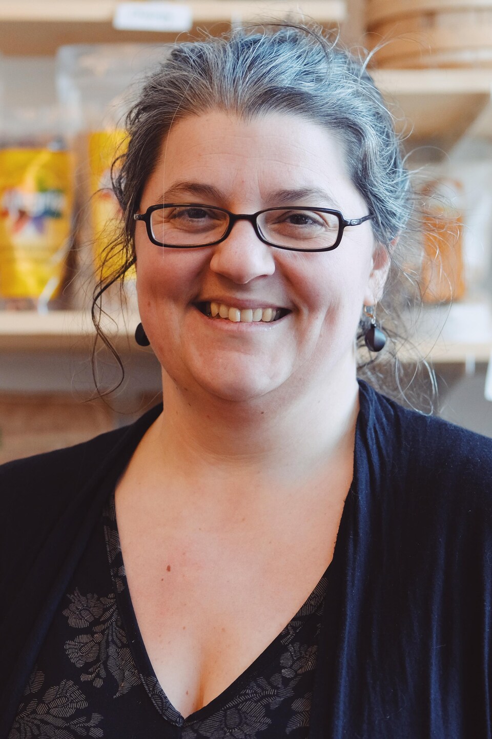 Annick Béland Morin est une femme aux cheveux grisonnants. Elle pose devant un étalage de produits locaux. 