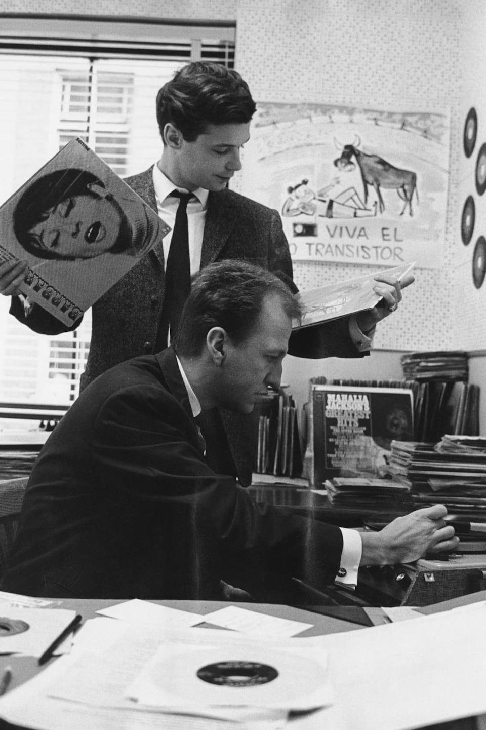 Dans un bureau, Jacques Boulanger, debout avec une pochette de disque dans chaque main, discute avec le réalisateur Gérard Binet qui, assis, fait fonctionner un tourne-disque.