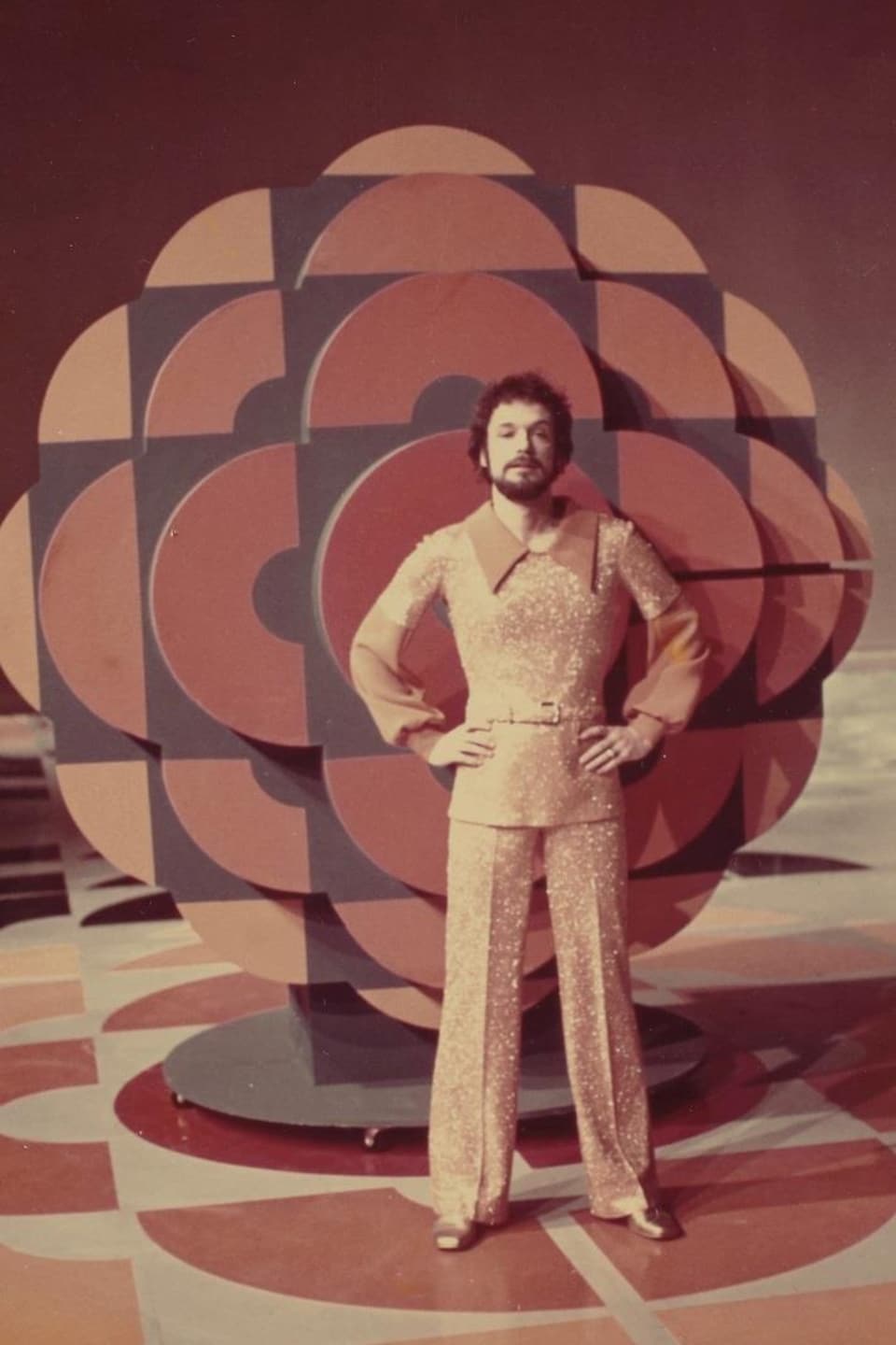 Jacques Boulanger, en tenue disco scintillante, posant devant un immense logo de Radio-Canada dans les mêmes tons.