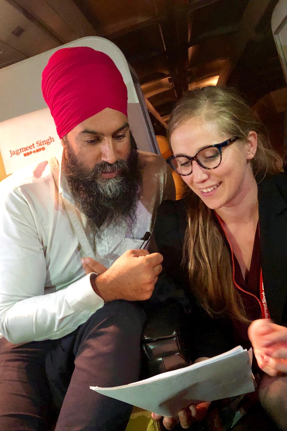 Jagmeet Singh est assis dans un avion, une jeune femme est agenouillée tout près de lui et lui montre un document.