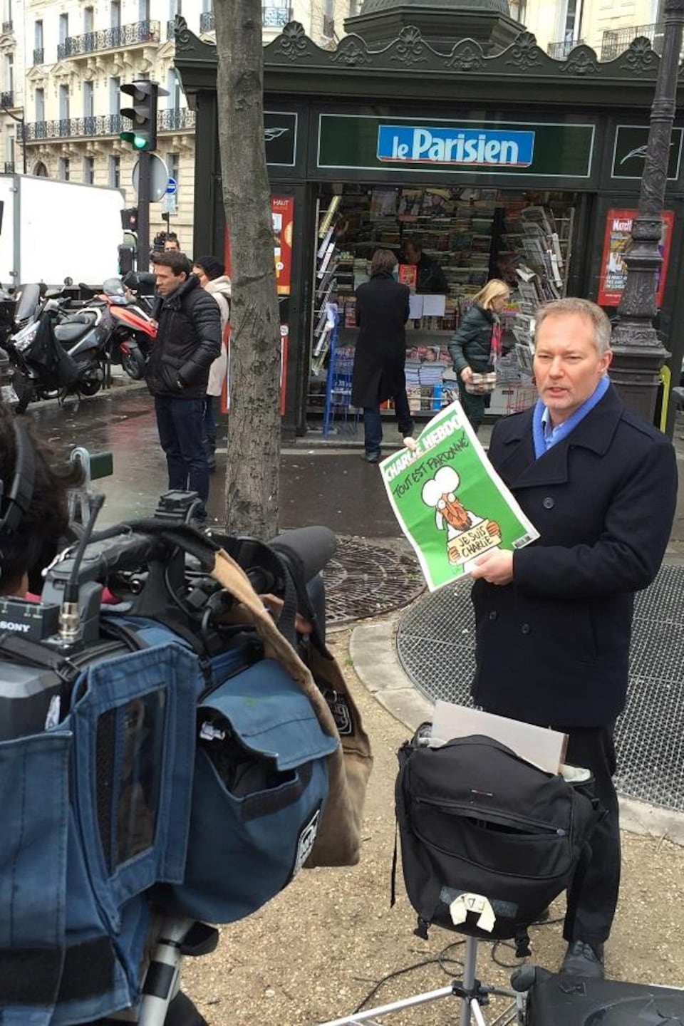 Le journaliste Jean-François Bélanger, correspondant à Paris, filmé par un caméraman, tenant entre ses mains un
exemplaire du Charlie Hebdo, devant un kiosque à journaux.
