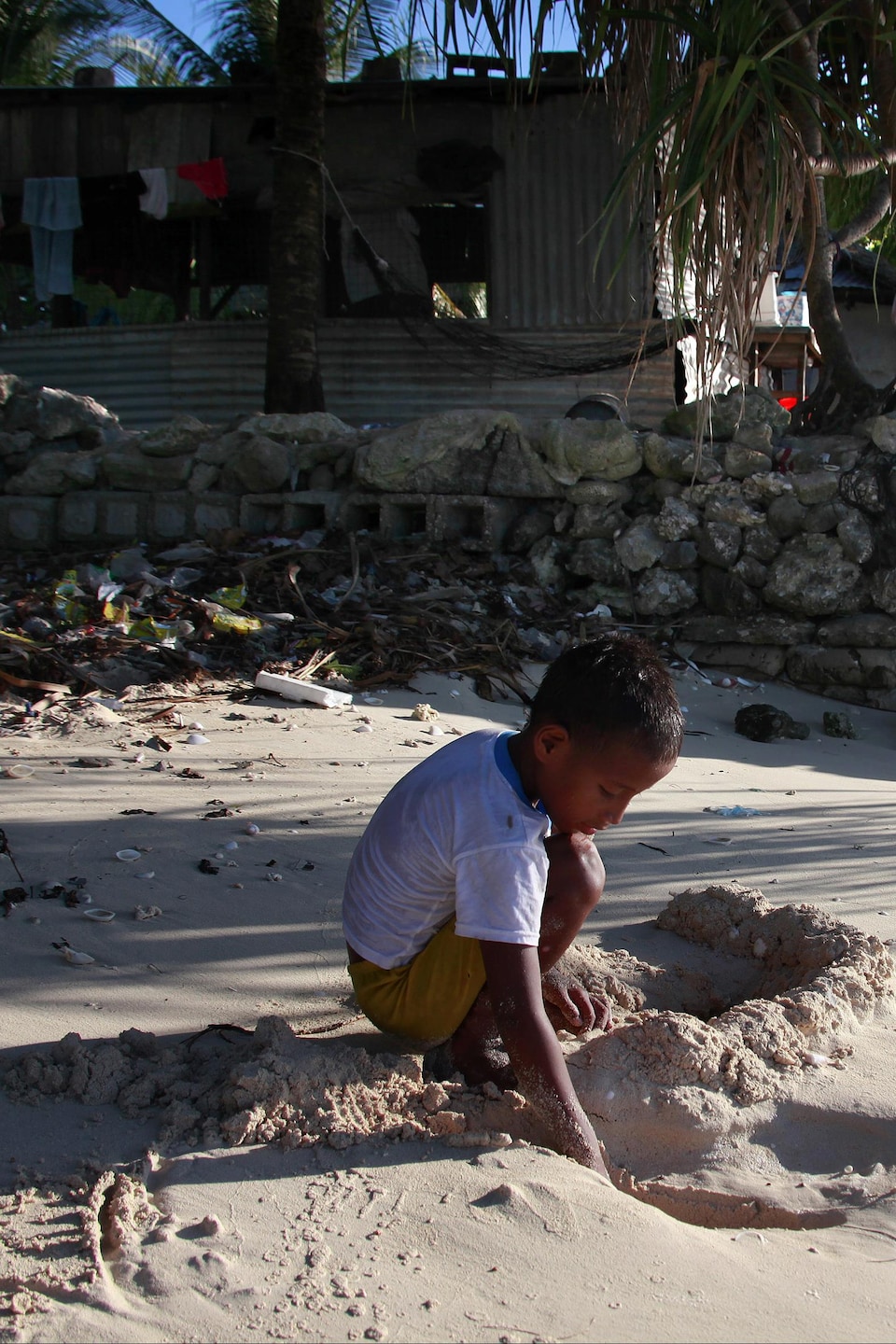 L'enfant joue dans le sable près de sa maison, qui est construite à côté de l'eau et entourée d'un muret de pierre.