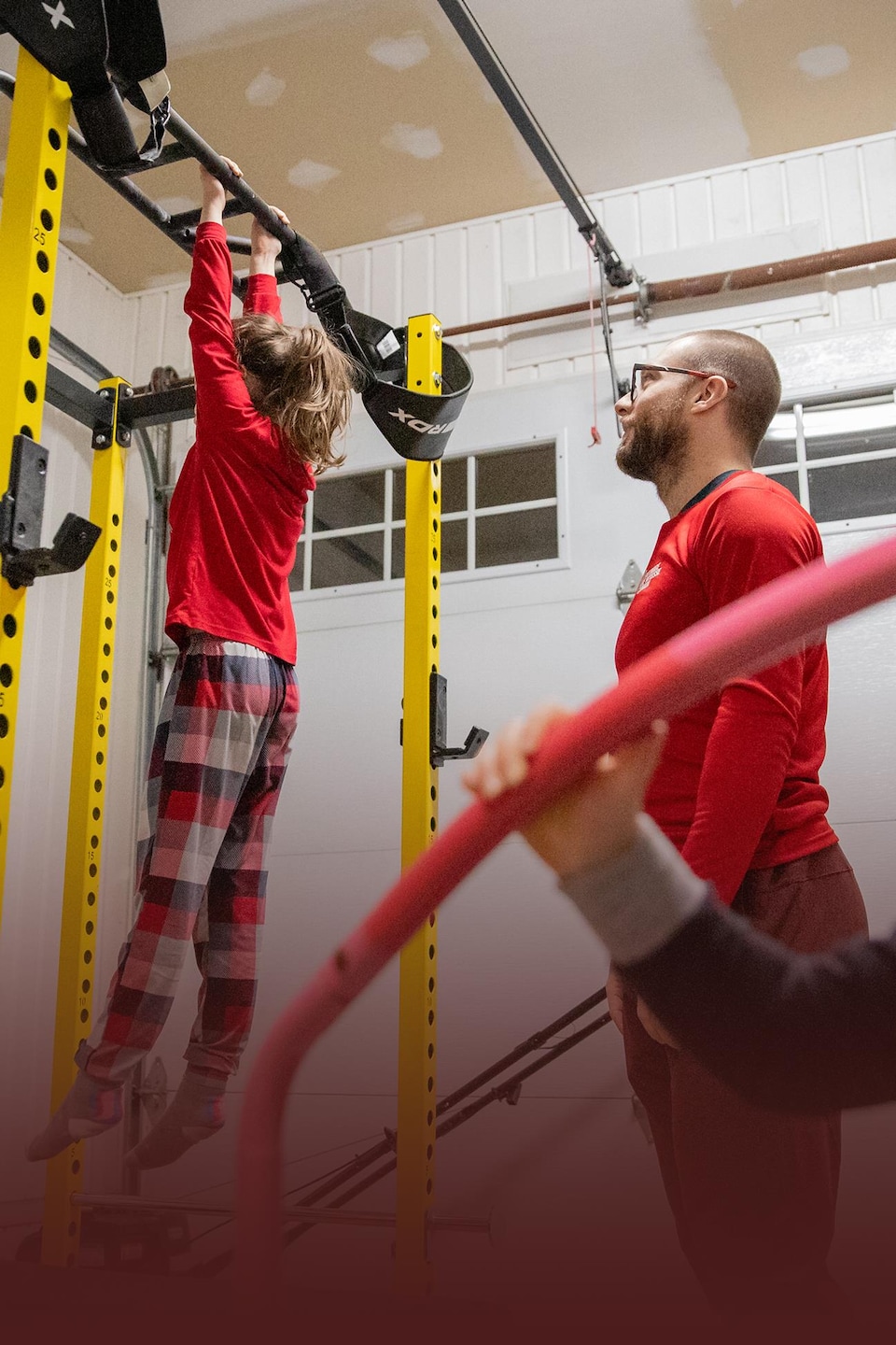 Un homme supervise une fille suspendue à une barre de traction dans un gymnase maison, tandis qu'un autre enfant est au premier plan.