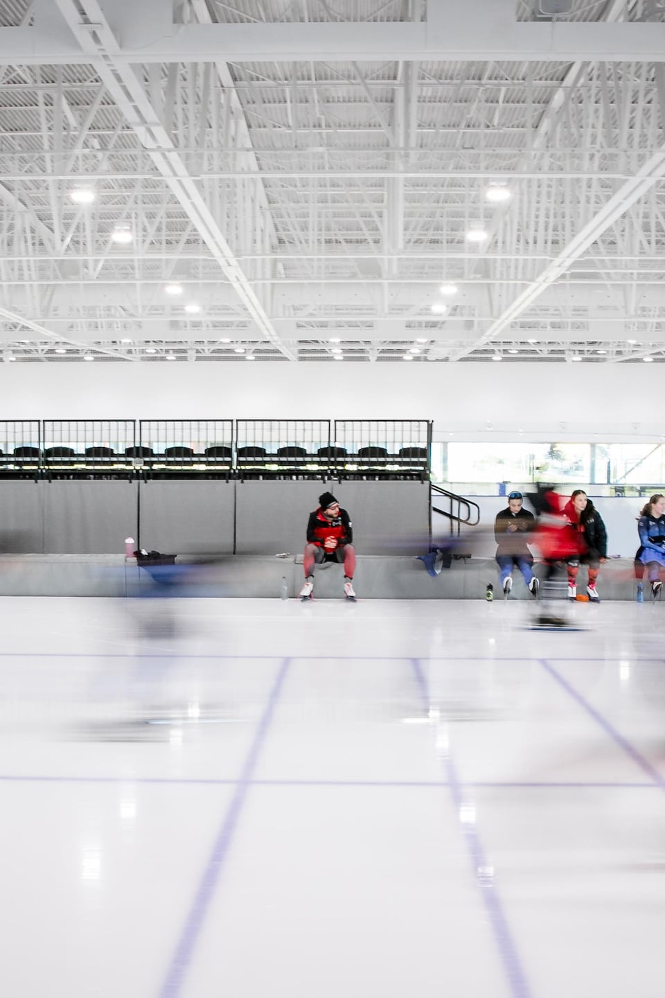 Vue d'ensemble d'une patinoire intérieure où plusieurs patineurs de vitesse s'entraînent, créant un effet de flou de mouvement. Laurent Dubreuil est assis sur le coussin en bordure du virage et les regarde passer.