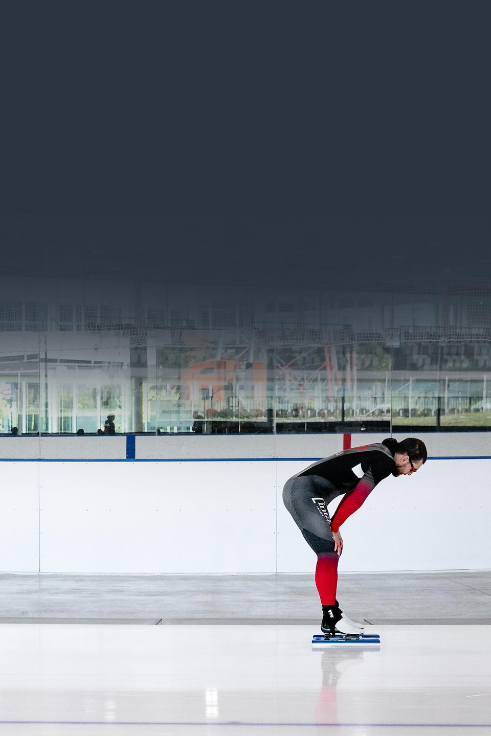 Un patineur de vitesse en combinaison noire et rouge se repose, penché vers l'avant sur une patinoire intérieure.