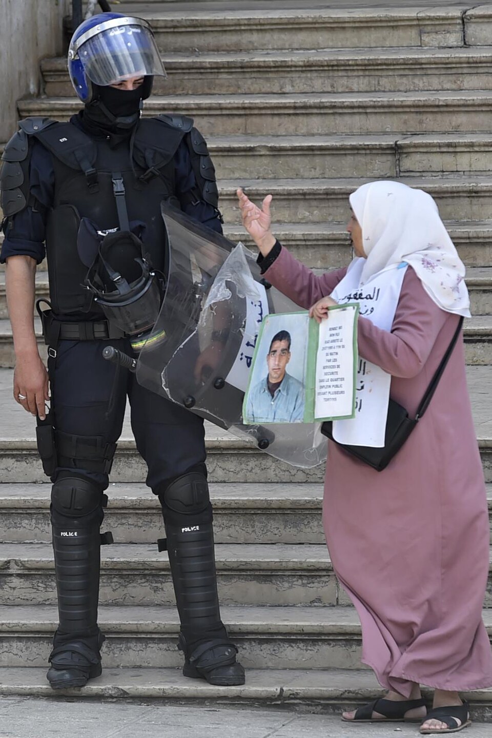 Une femme tient une pancarte sur laquelle on voit un jeune homme et un court texte devant un policier casqué, armé et portant des équipements de protection.