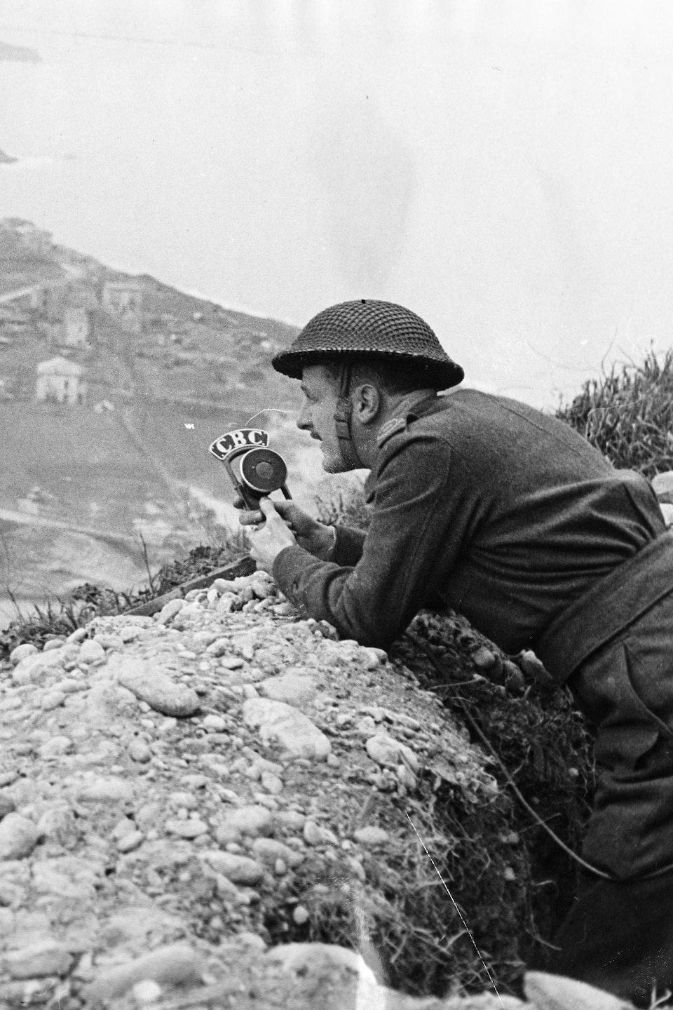 Juché sur une butte sur le front italien, le correspondant de guerre Marcel Ouimet, portant un casque militaire, enregistre un reportage un micro à la main.