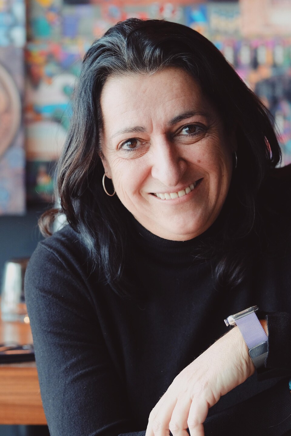 Marie-Chantal Lepage est assise sur une chaise et pose, souriante, pour la caméra devant un tableau coloré et contemporain.
