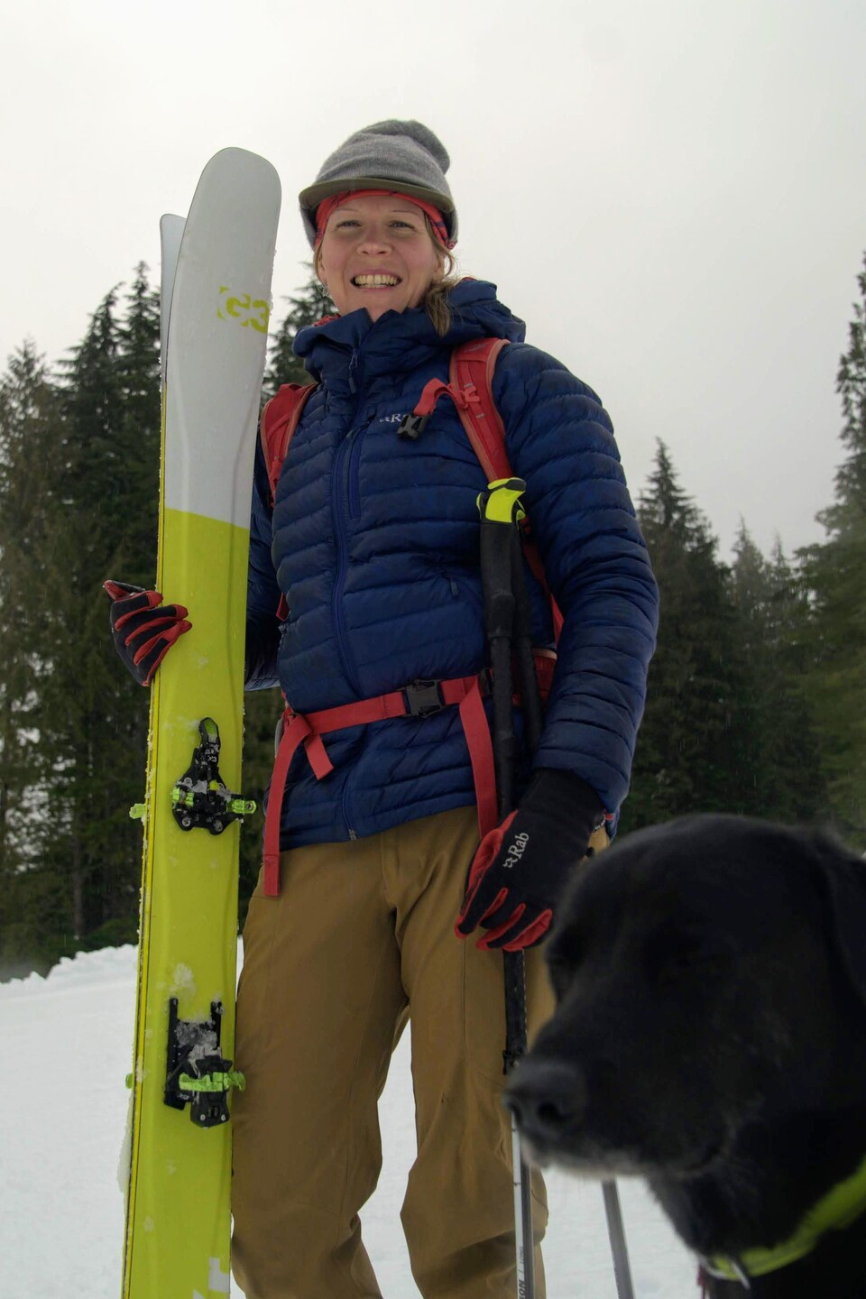 Une femme dans la trentaine est debout avec ses skis alpins à la main devant une forêt et de la neige avec un chien noir à ses pieds. 