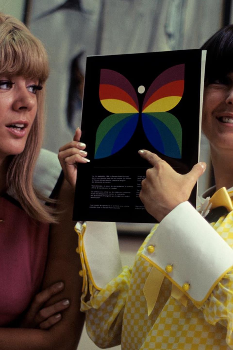 Dans un bureau, les comédiennes Denise Filiatrault et Dominique Michel, avec le papillon multicolore de Radio-Canada entre les mains.