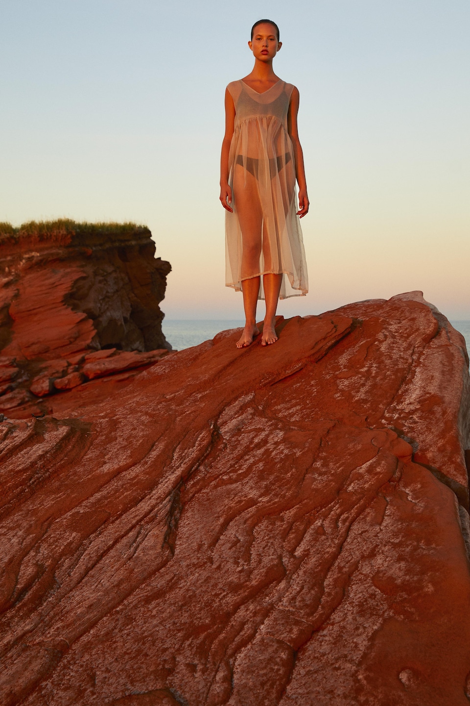 Une mannequin en robe diaphane se tient sur un rocher, au coucher de soleil.