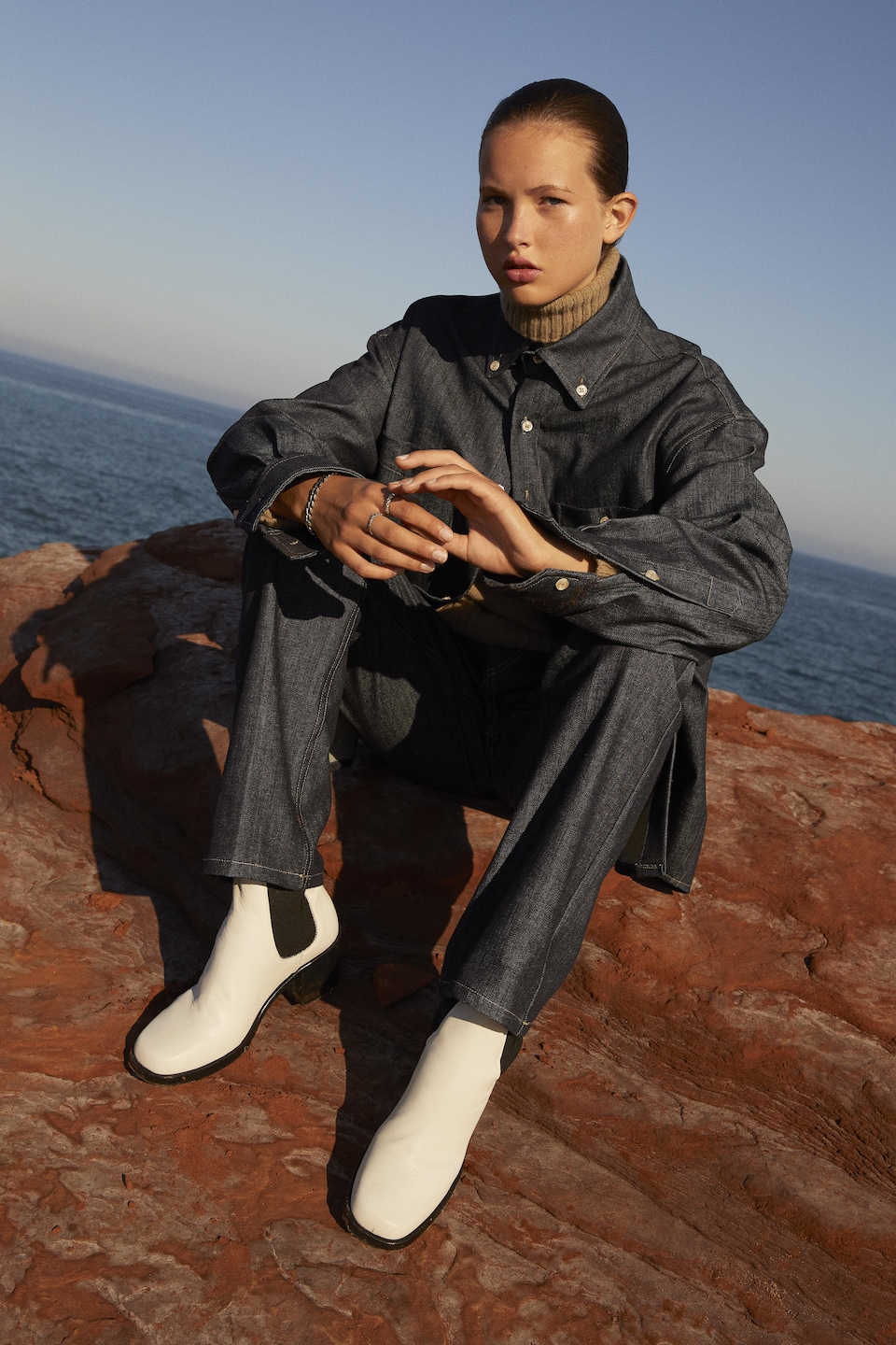 La mannequin est assise sur des rochers, dos à la mer, et porte des bottes blanches en cuir.