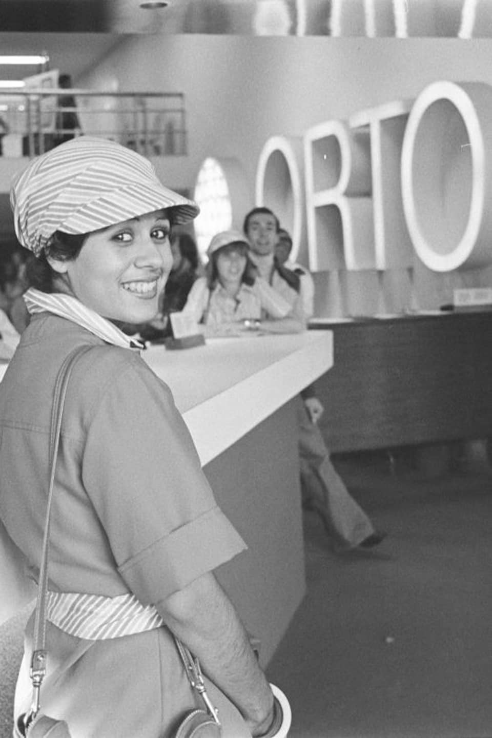 Une hôtesse sourit à la caméra à l'entrée de la salle de l'Organisme de radio-télévision des Olympiques (ORTO).