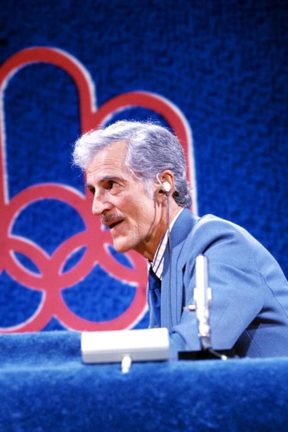 En studio, le journaliste René Lecavalier seul au micro. Derrière lui, le logo des Jeux olympiques de Montréal.