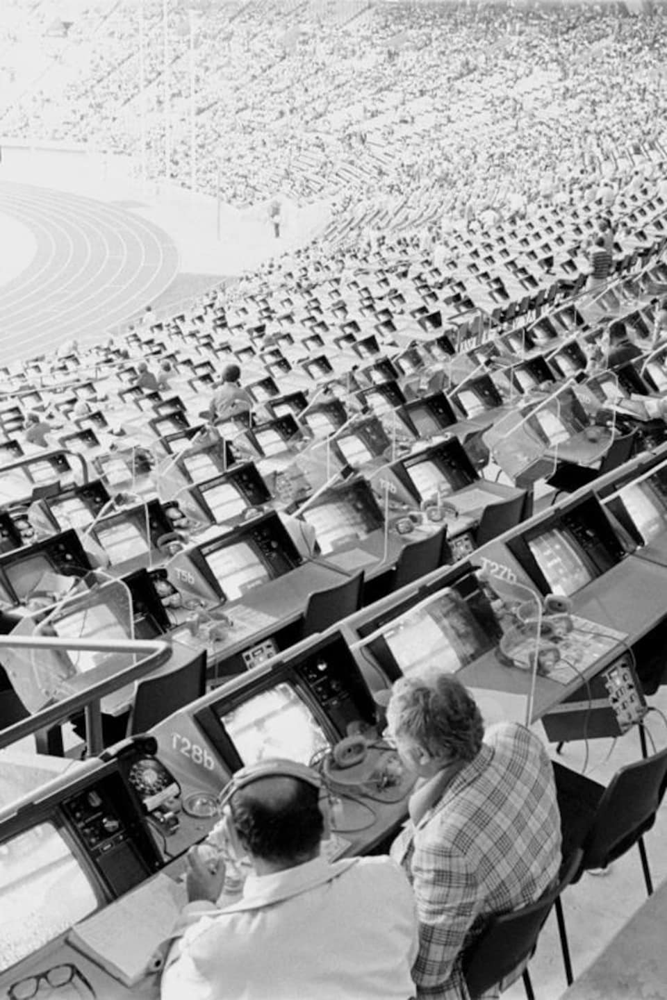 Vue générale des gradins et des tribunes du stade olympique avec quelques commentateurs assis à leur consolette.