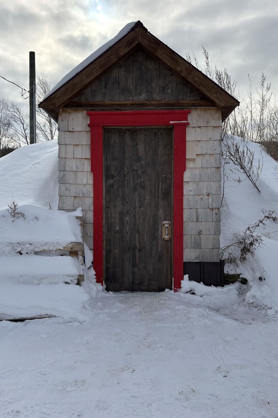 Une petite cabane en bardeaux de cèdre est adossée à une butte de neige.