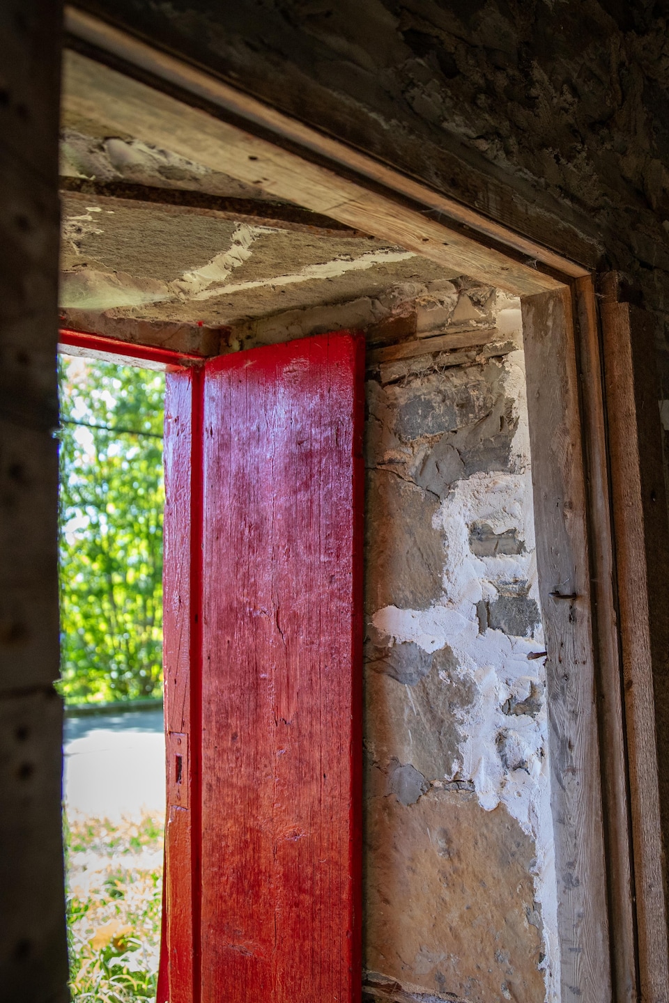 Vue depuis l’intérieur d'un caveau en pierre, une porte en bois peinte en rouge s'ouvre sur un paysage verdoyant et ensoleillé.