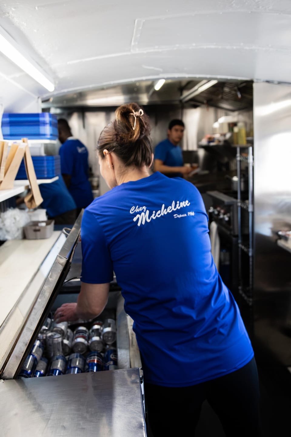 Vue de dos, une employée portant un t-shirt bleu marqué du logo « Chez Micheline » prend une canette dans un réfrigérateur.