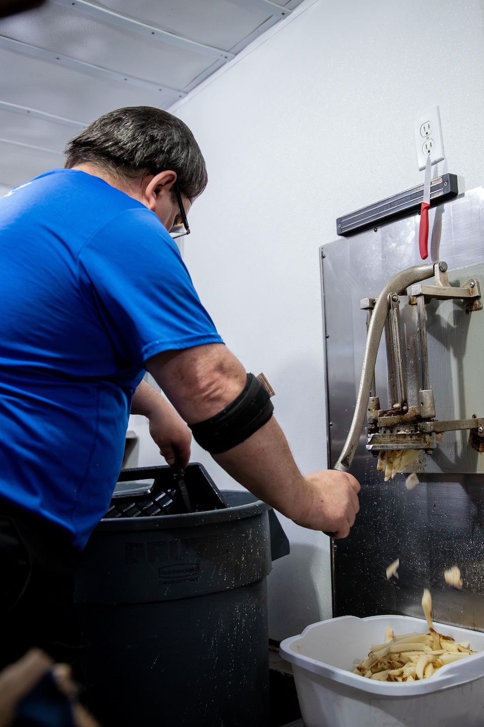 Un employé utilise un coupe-frites manuel fixé au mur pour découper des pommes de terre au-dessus d'un bac.