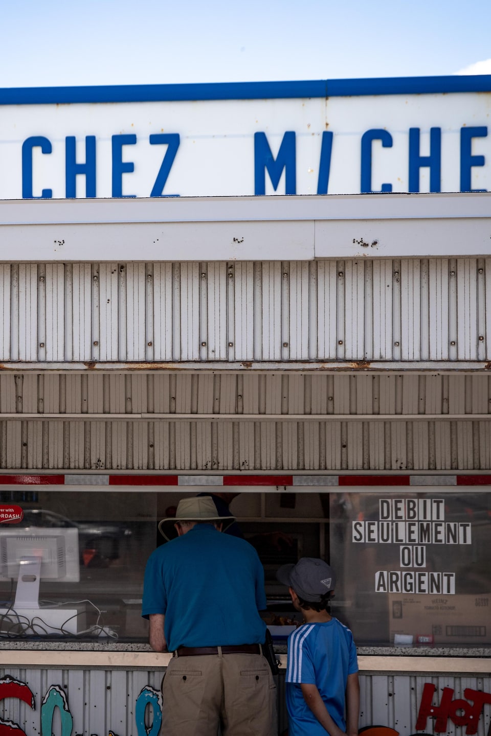 Deux clients passent une commande au comptoir de la cantine « Chez Micheline », aux côtés des affiches « Recevez ici » et « Débit seulement ou argent ».