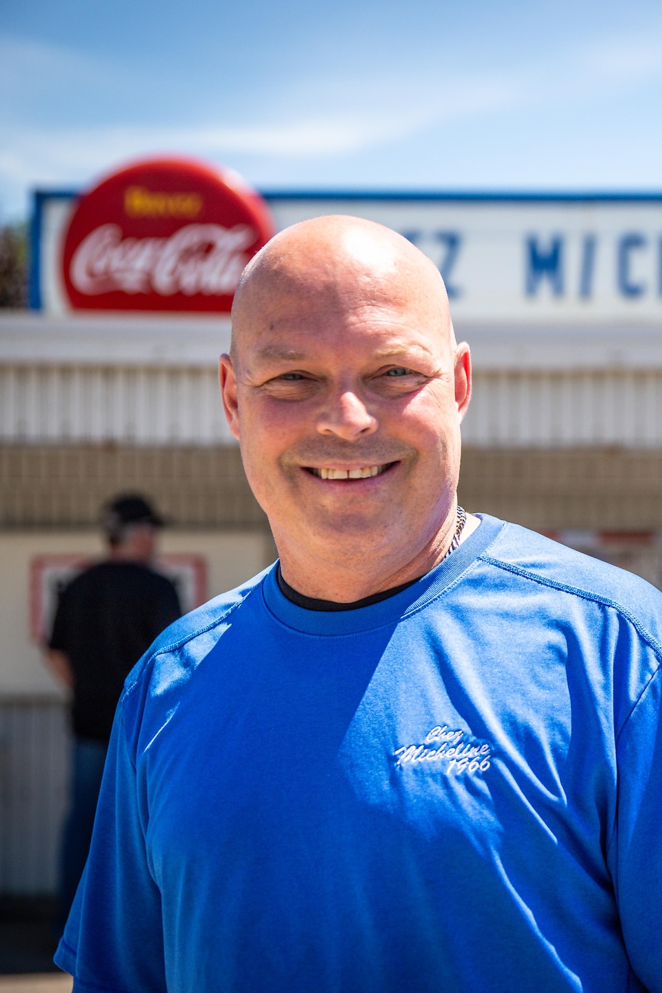 Un homme chauve et souriant vêtu d'un t-shirt bleu pose devant le commerce « Chez Micheline ».  