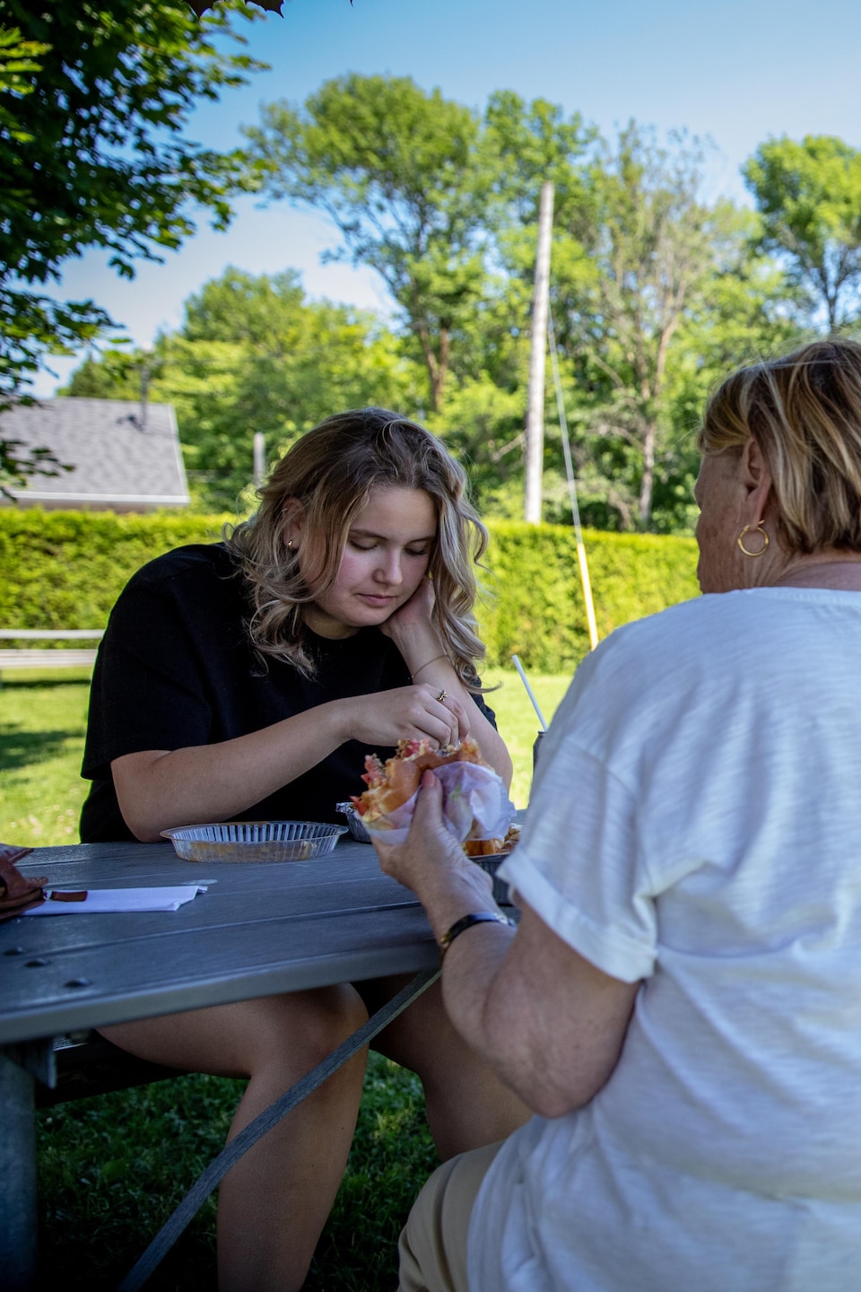 Deux personnes mangent à une table de pique-nique dans un parc avec des jeux pour enfants en arrière-plan.
