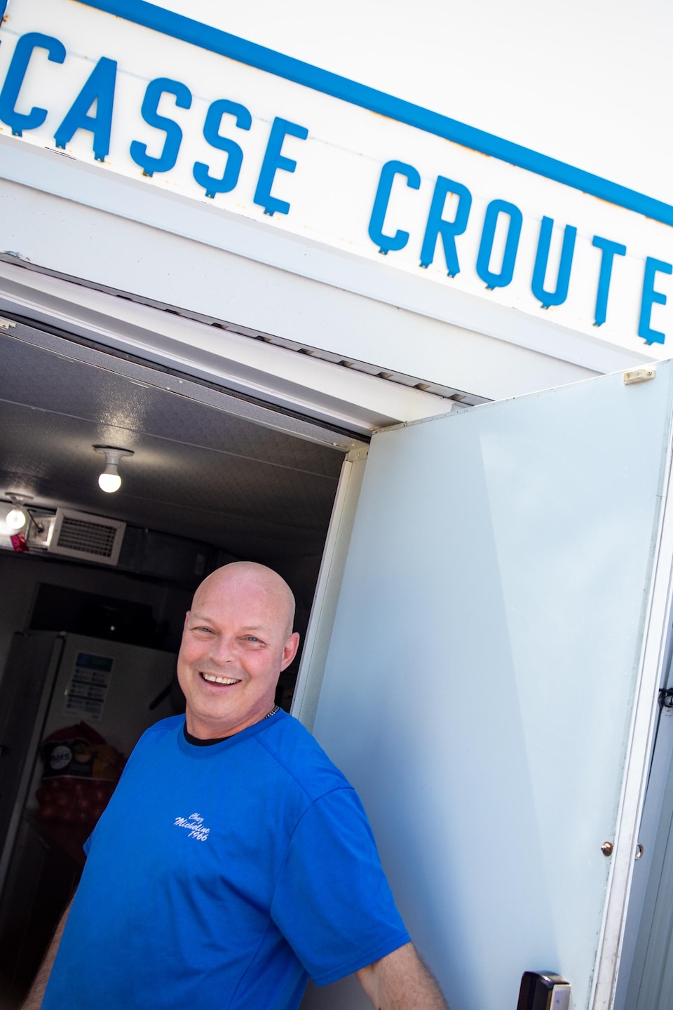 Un homme souriant en t-shirt bleu se tient à la porte d'un bâtiment sous l'enseigne « Casse Croûte ».