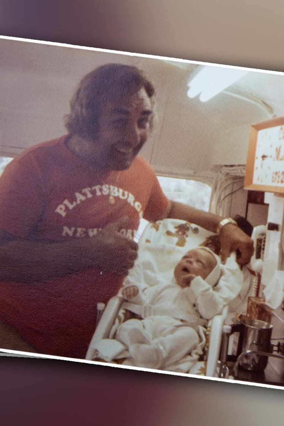 Un homme souriant en chandail rouge pose à côté d’un bébé installé dans une chaise haute.