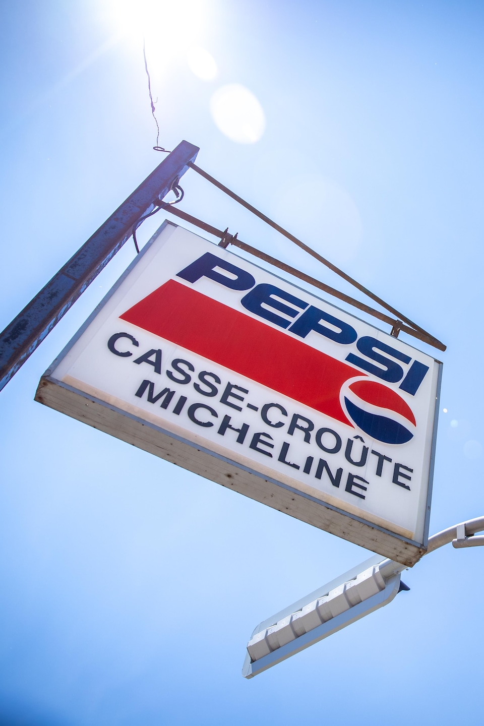 Météo ensoleillée avec une enseigne « Pepsi Casse-croûte Micheline » suspendue à une structure métallique.