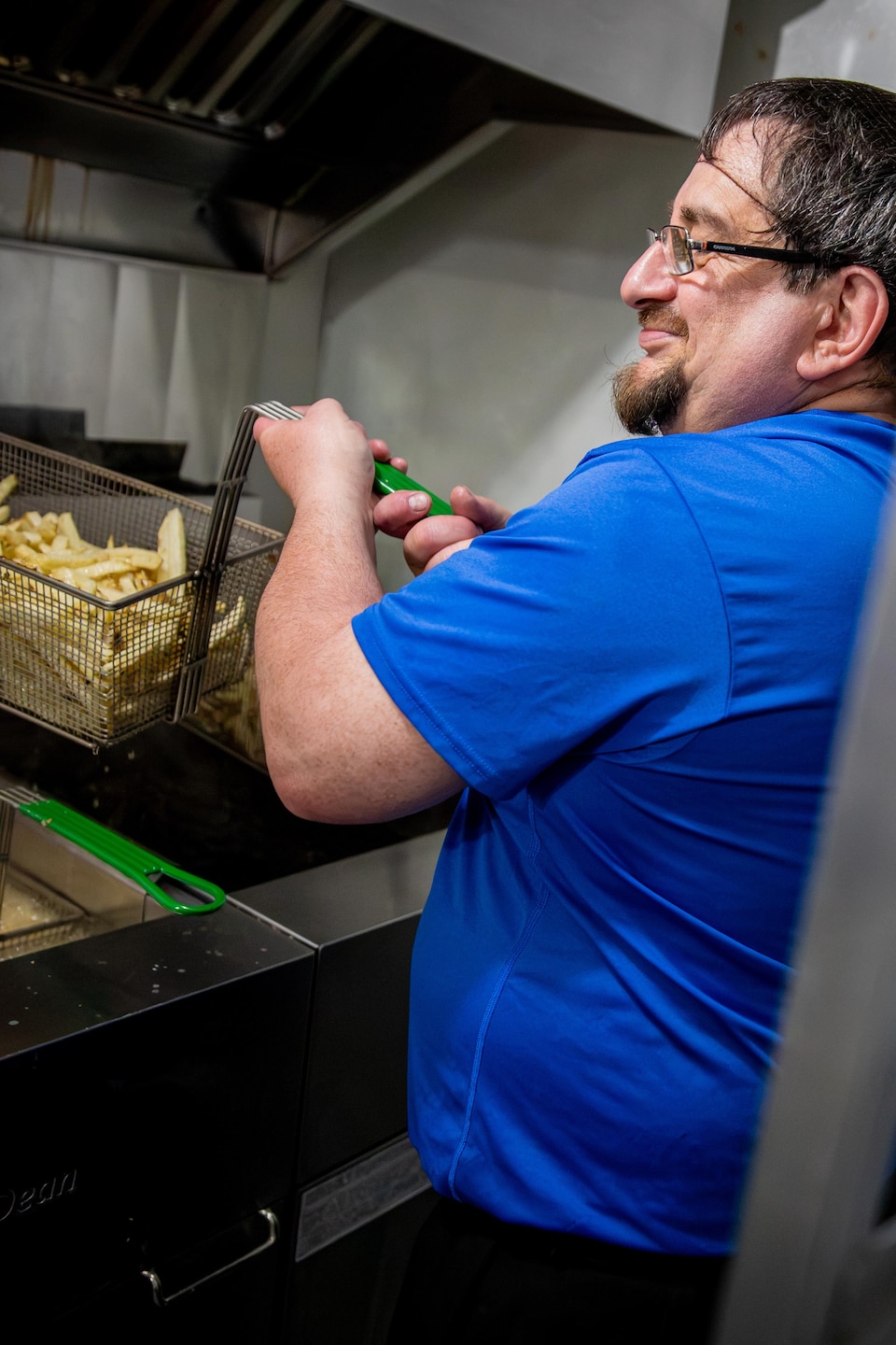 Un employé en t-shirt bleu retire un panier de frites d'une friteuse dans une cuisine.