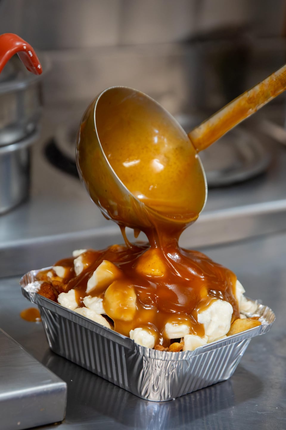 Une louche verse de la sauce brune chaude sur une poutine garnie de fromage en grain.