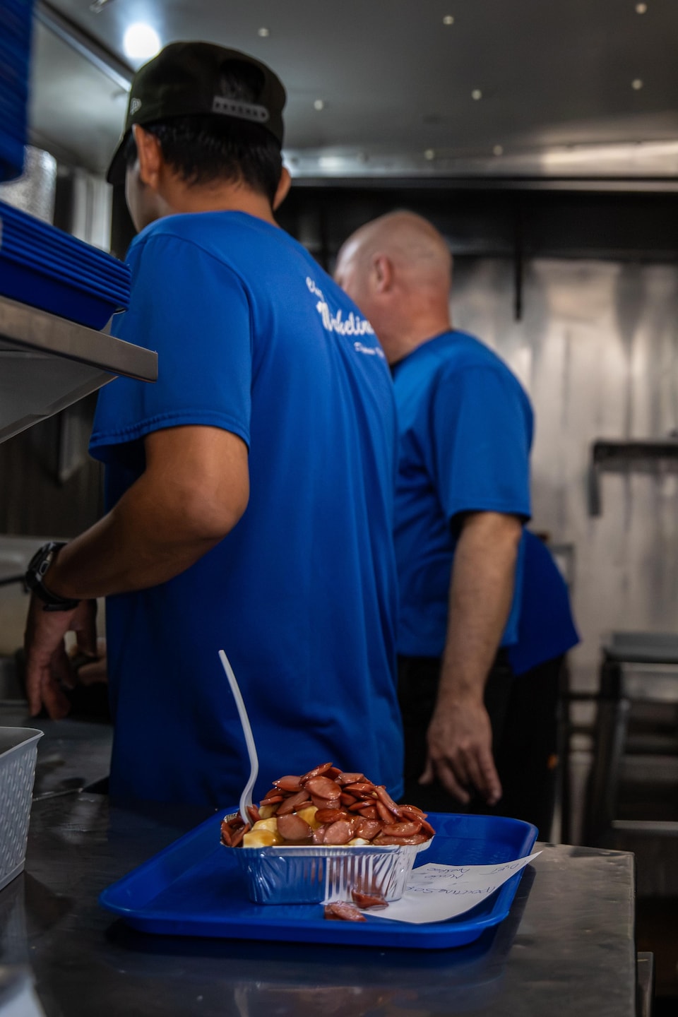 Trois employés en t-shirt bleu préparent de la nourriture en cuisine, devant une poutine garnie de saucisses servie sur un plateau.