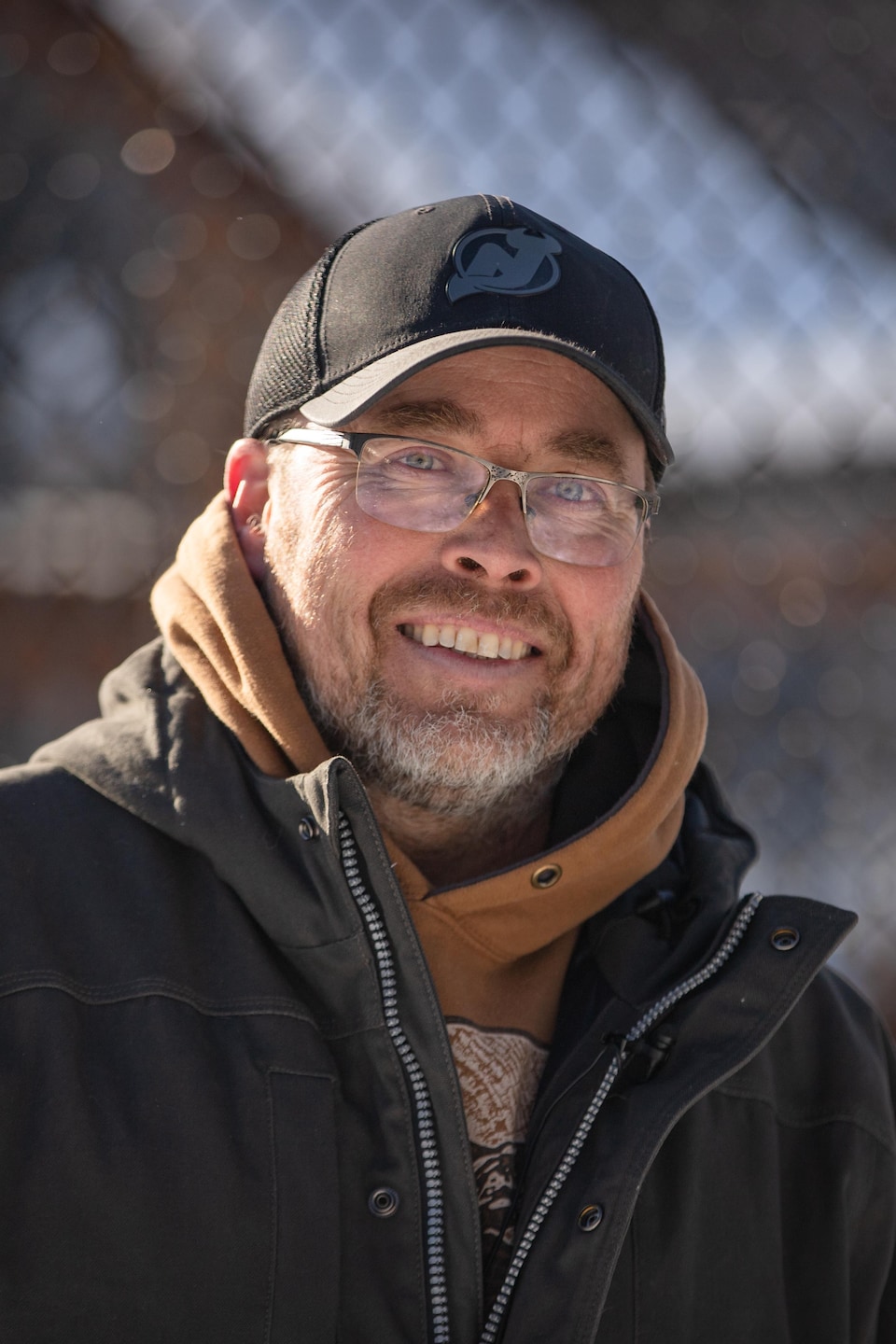 Un homme souriant portant une casquette noire, des lunettes et un manteau d'hiver pose devant une clôture grillagée.