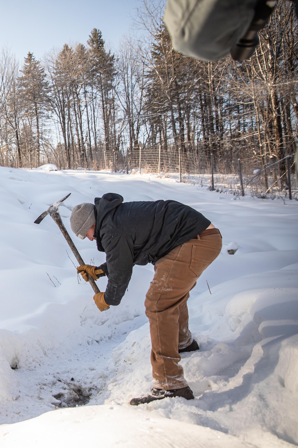 Un homme penché utilise un pic pour casser la glace d'un ruisseau dans un enclos enneigé.
