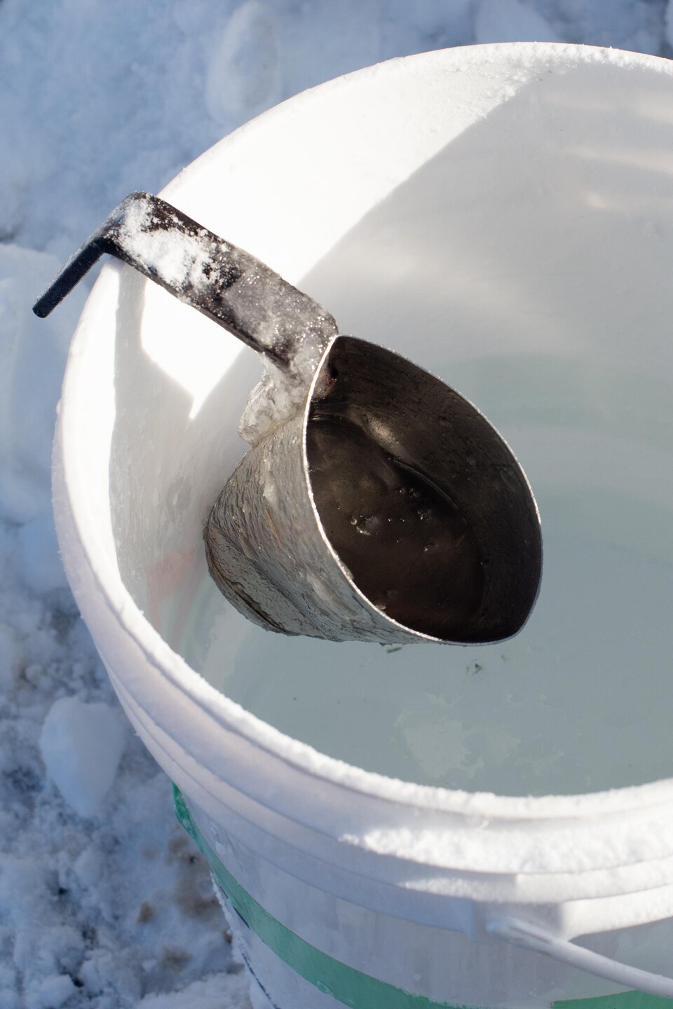 La louche en métal est accrochée sur le bord d'un sceau d'eau, en apparence légèrement glacée.