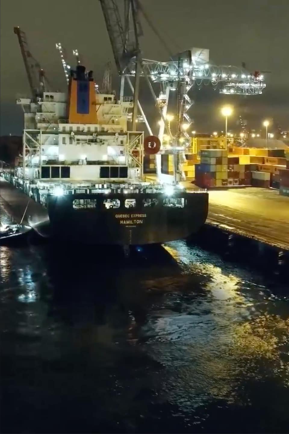 De petits bateaux poussent un porte-conteneurs vers un quai au port de Montréal en pleine nuit.