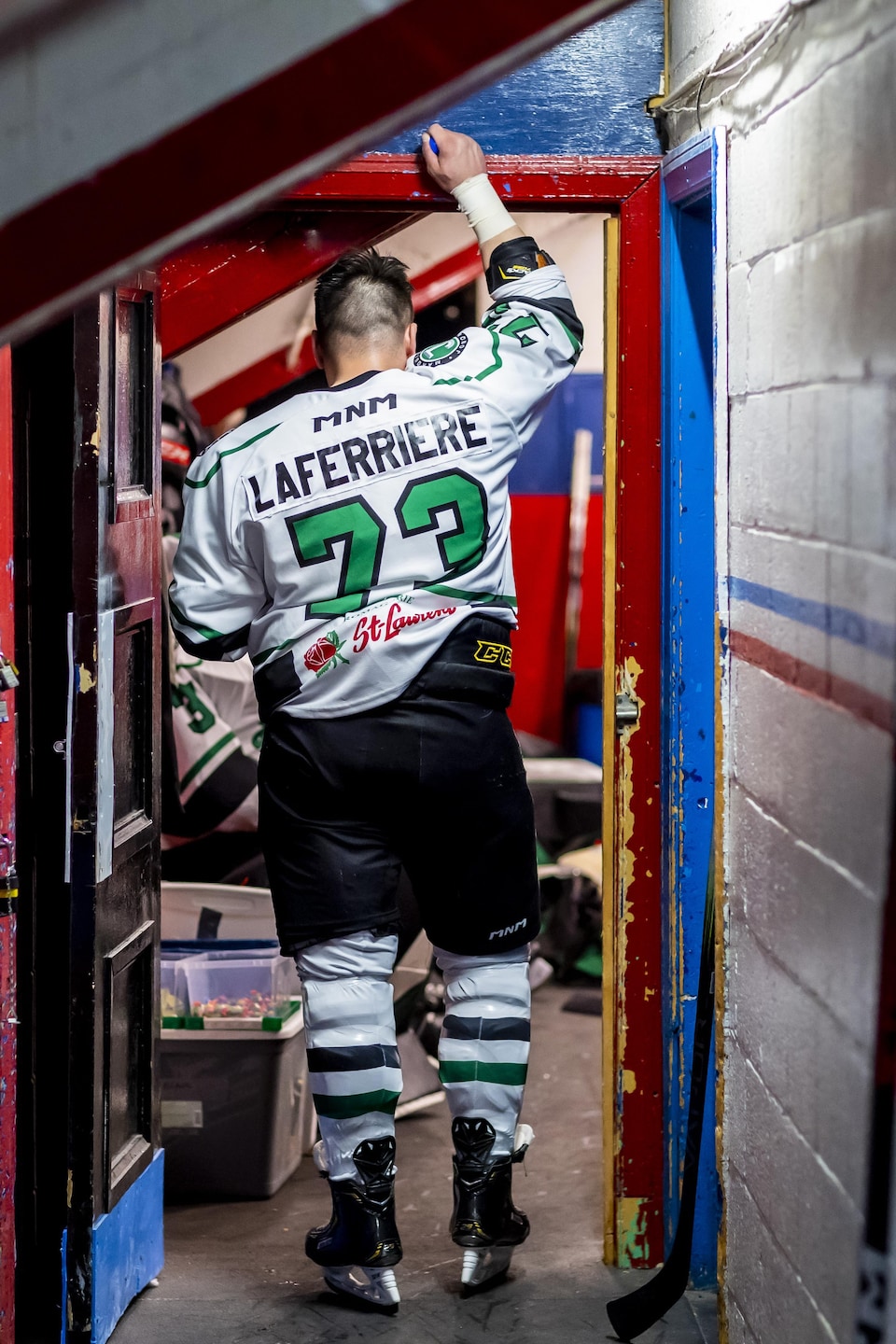 Un joueur de hockey en blanc debout, de dos, devant le vestiaire de son équipe. 