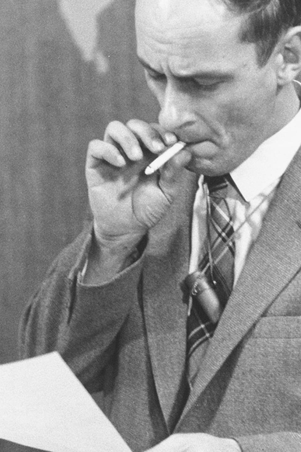 Devant une carte du monde, l'animateur René Lévesque tire sur sa cigarette, tenant une feuille dans l'autre main.