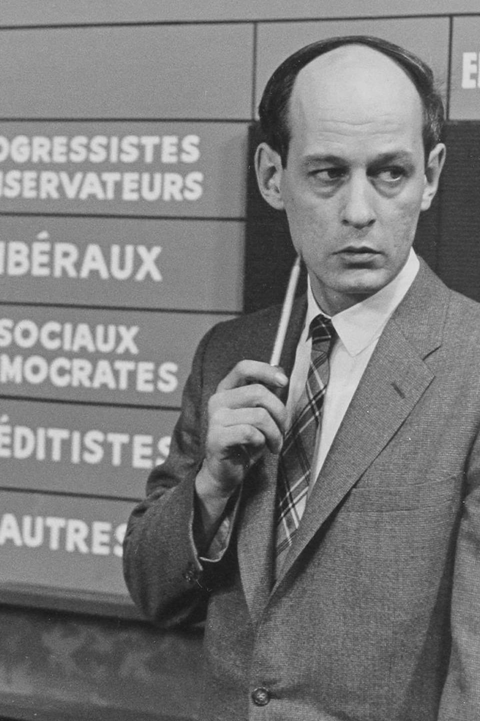 René Lévesque debout, baguette à la main, devant un tableau des résultats électoraux.
