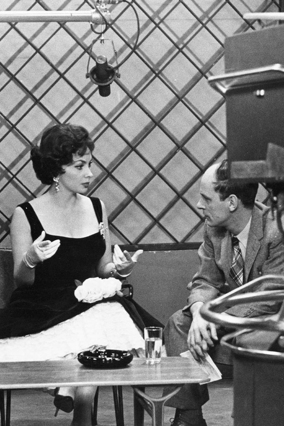 Dans un studio de télévision, le journaliste René Lévesque écoute l'actrice italienne Gina Lollobrigida. Un caméraman non identifié de dos filme l'entrevue, la silhouette d'une deuxième caméra au premier plan à gauche ainsi qu'un microphone au bout d'une perche complètent le décor.