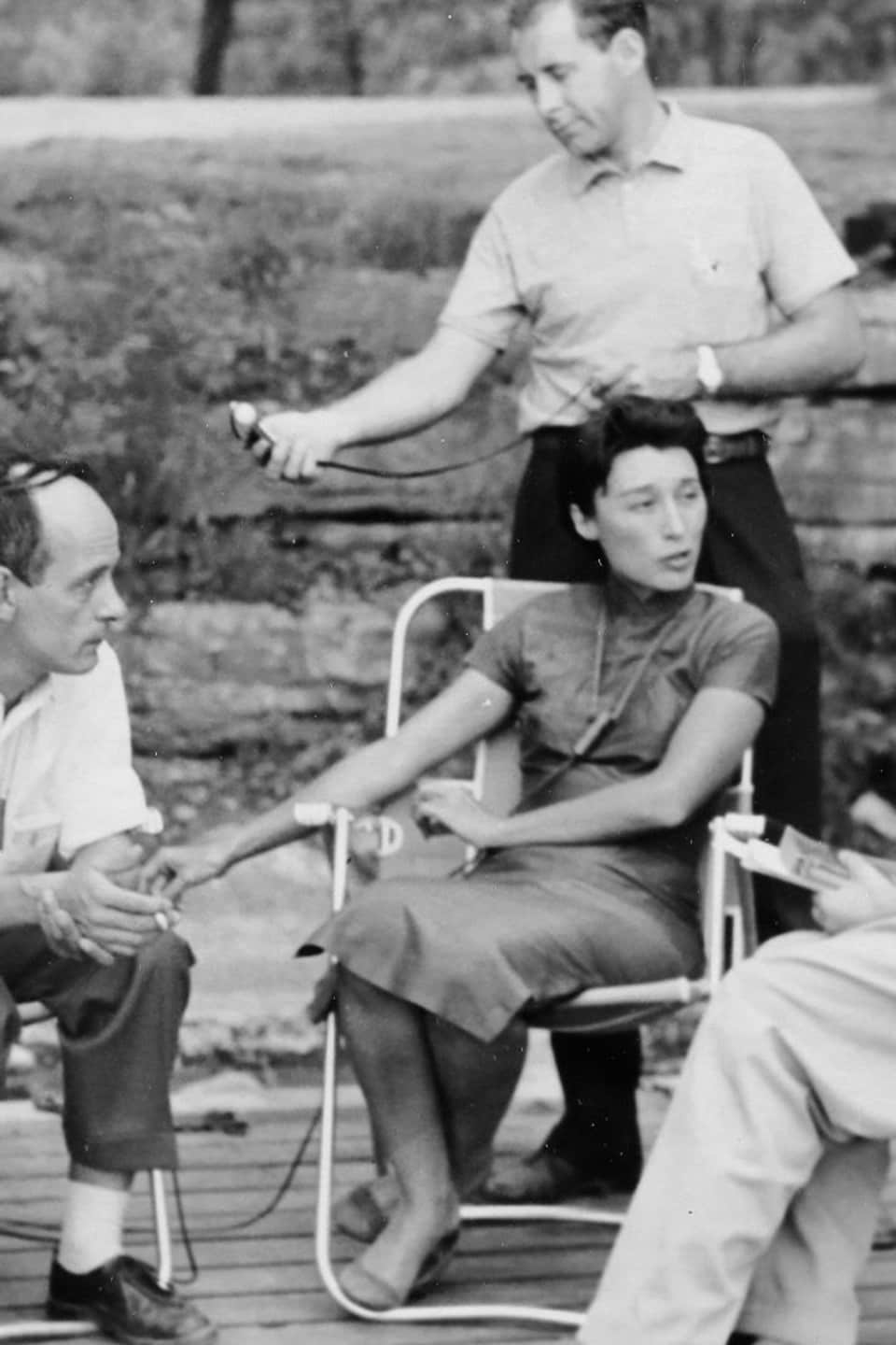 René Lévesque et Han Suyin assis à l'extérieur sur des chaises de jardin. D'autres personnes sont autour d'eux, dont un preneur de son. 