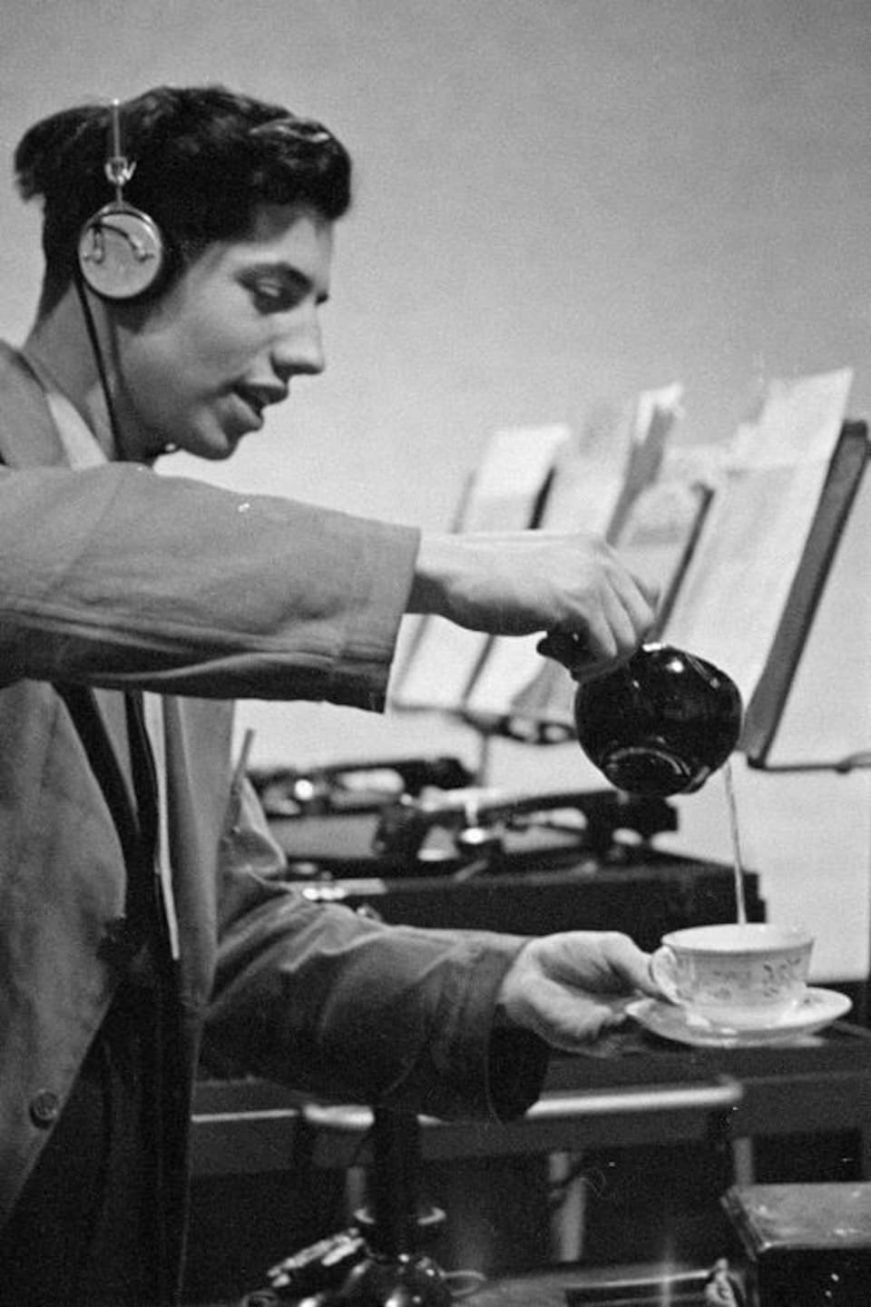 Dans un studio de radio, derrière un microphone sur pied, le bruiteur Marcel Giguère produit le son de l'eau versé dans une tasse.