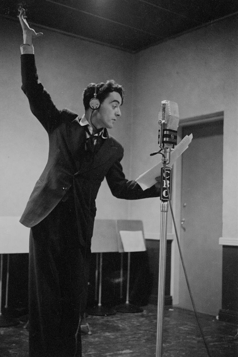 Dans un studio de radio, derrière un microphone sur pied, le réalisateur Guy Mauffette s'exprimant avec de grands gestes.