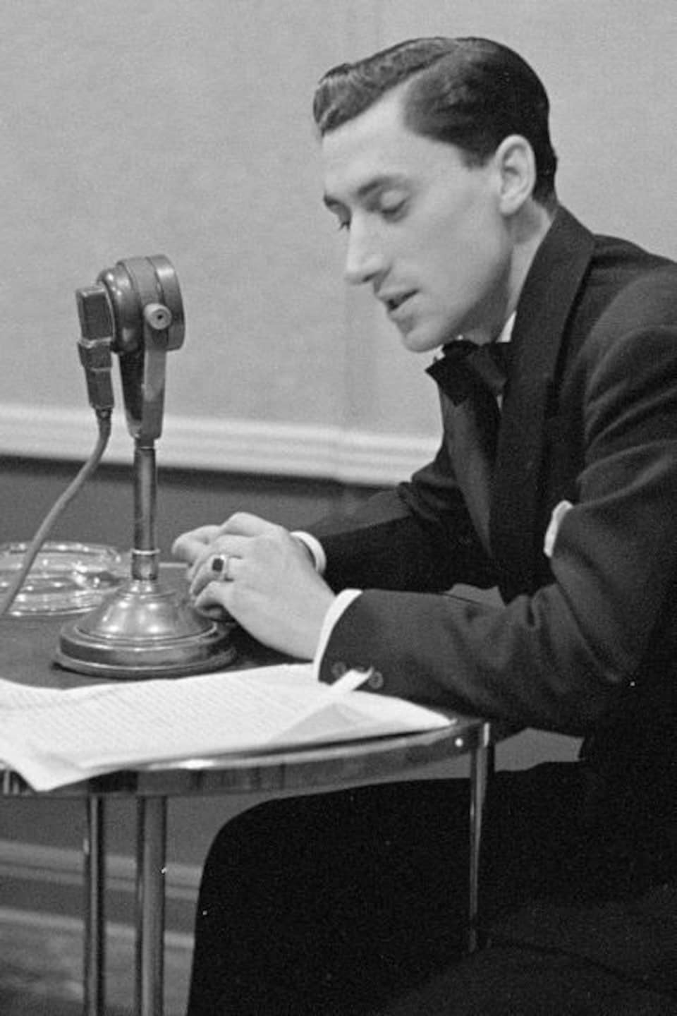 Dans un studio de radio, l'annonceur Roger Baulu installé à une petite table surmontée d'un micro sur pied.