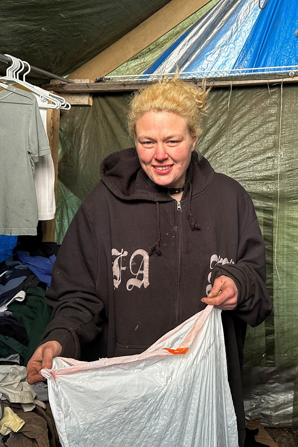 Sheena Derdak est debout à l'intérieur d'une cabane où des vêtements sont accrochés sur des cintres et tient un sac de déchets blanc qui contient des dons.