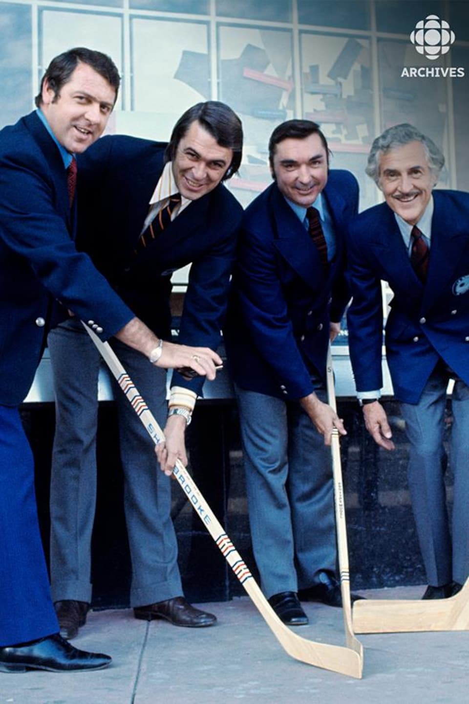À l'extérieur, debout devant l'édifice du Forum de Montréal, l'analyste Gilles Tremblay et les commentateurs sportifs
Richard Garneau, Lionel Duval et René Lecavalier avec des bâtons de hockey dans les mains.